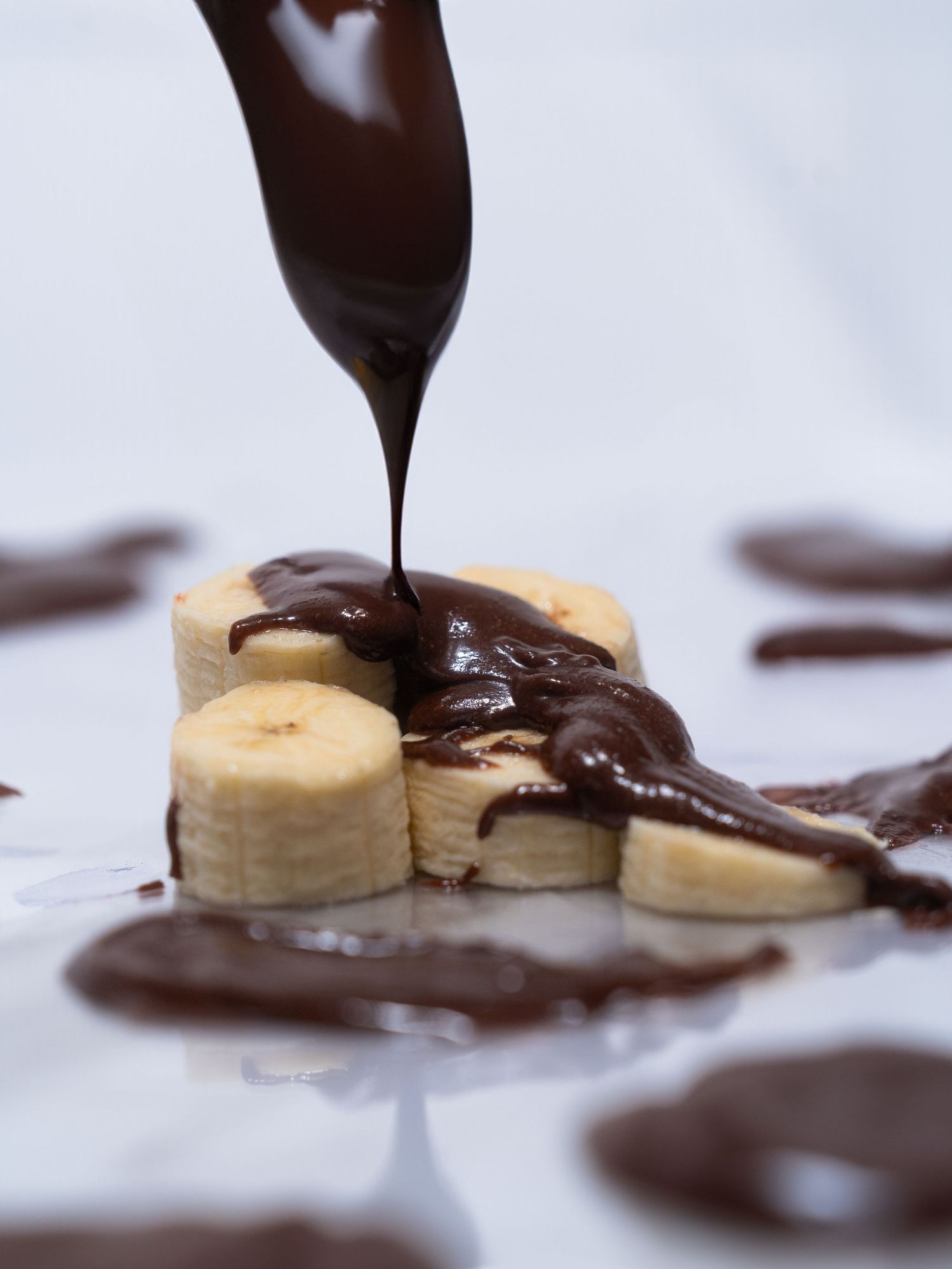 Melted chocolate being poured over sliced bananas on a white surface.