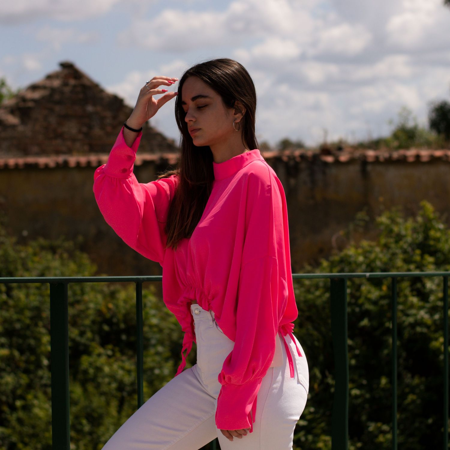 Woman in white dress and bright pink jacket posing against stone wall on sunny day.