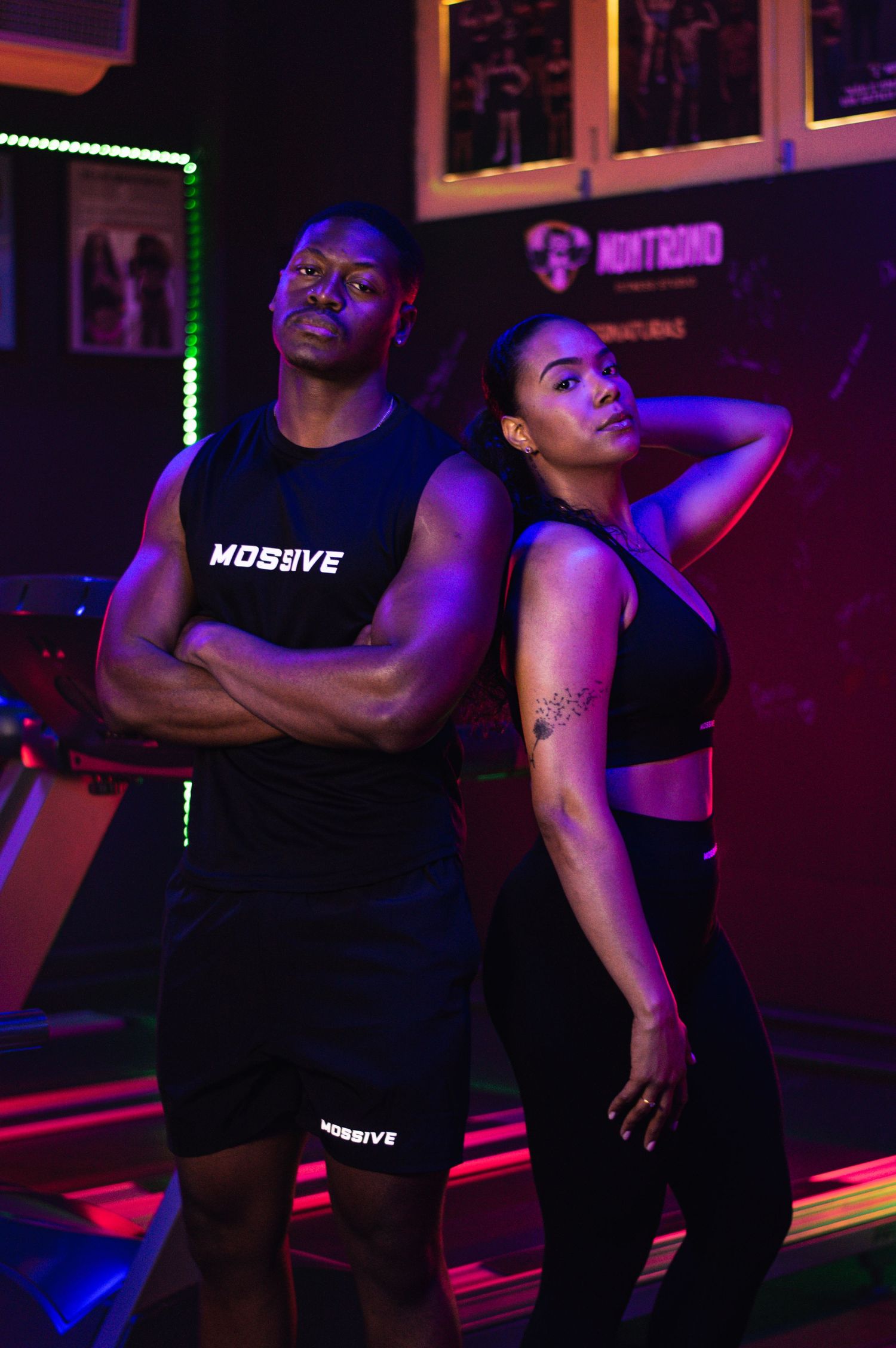 Two athletes in black MOSBVE workout gear posing in gym with neon lighting.