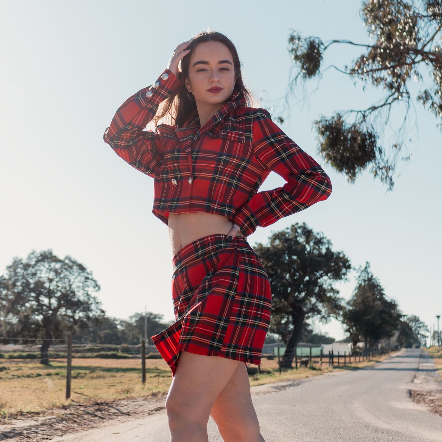 Model in red plaid outfit posing on rural road with eucalyptus trees in background.