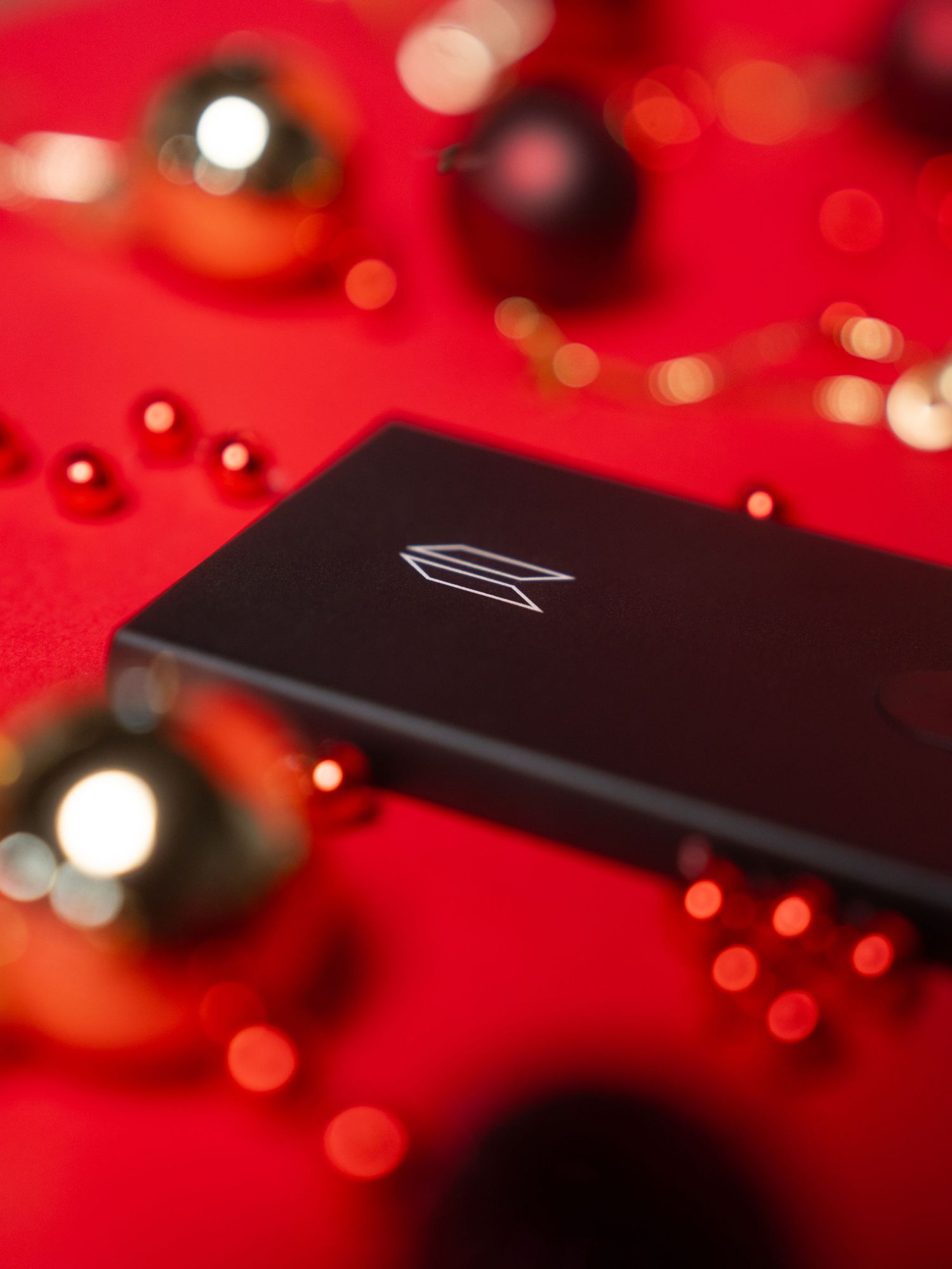 A sleek black electronic device against a red background with holiday ornaments and bokeh lights.