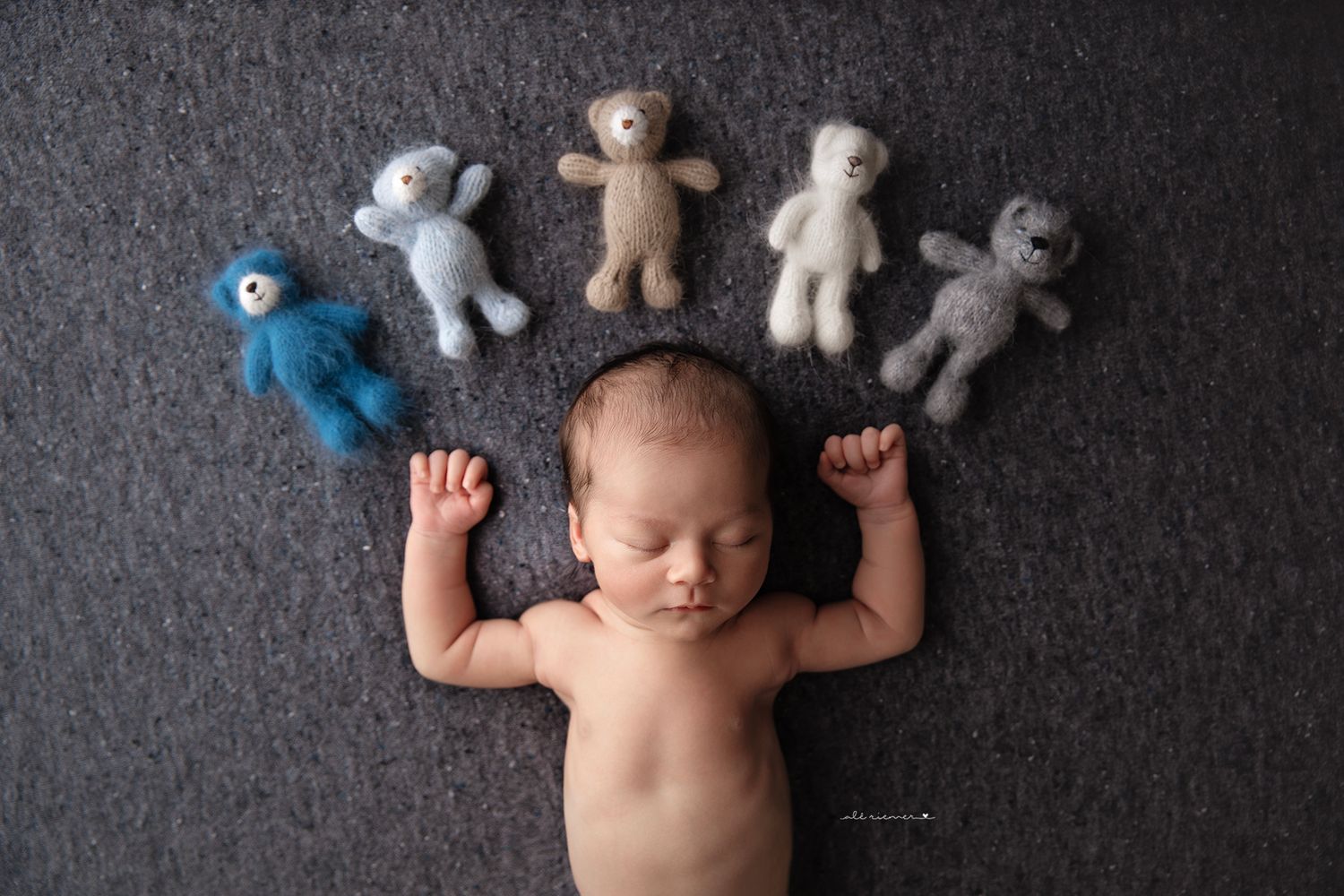 A sleeping newborn on grey fabric surrounded by small stuffed animal toys.