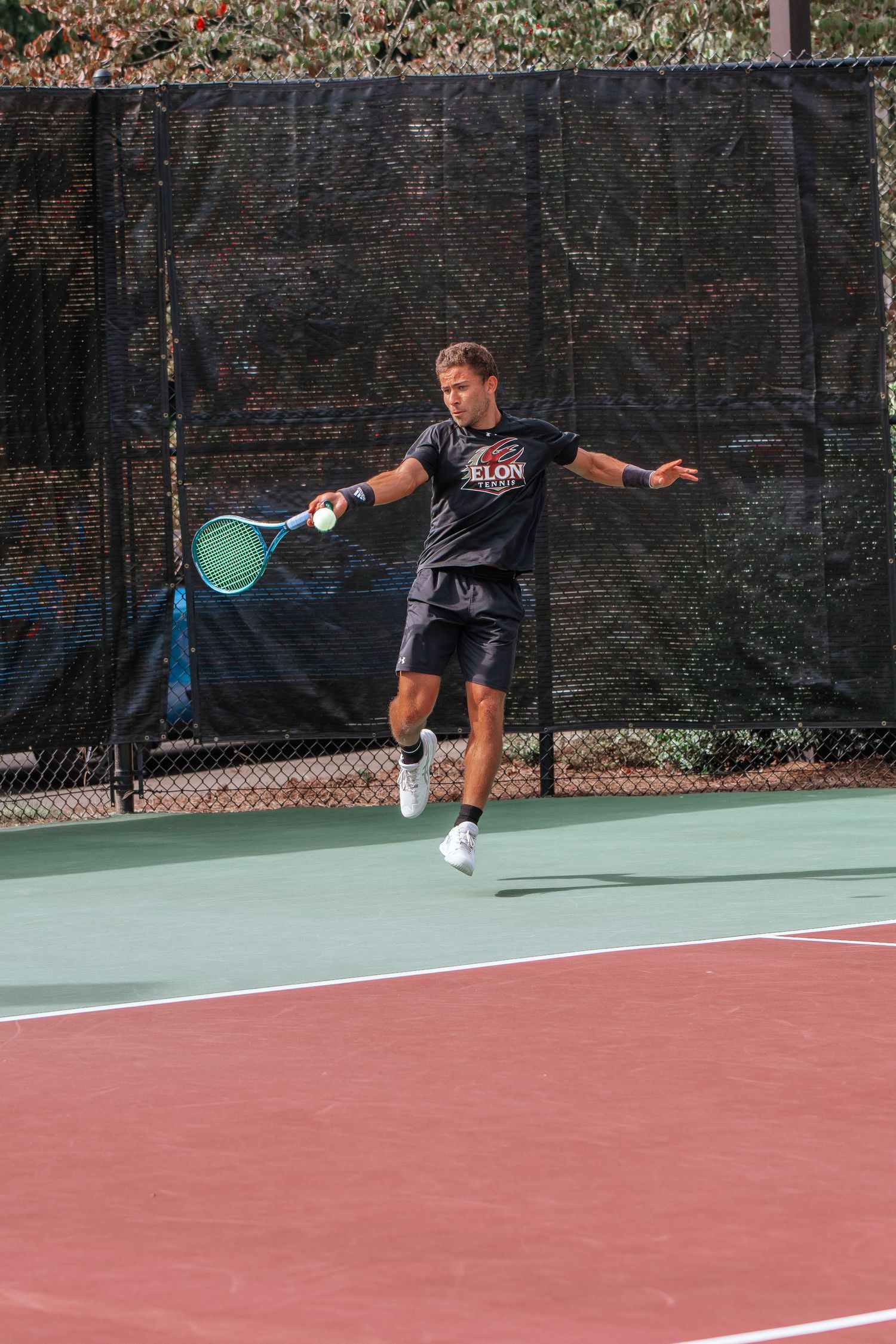 A tennis player mid-swing on an outdoor court with a multicolored surface.
