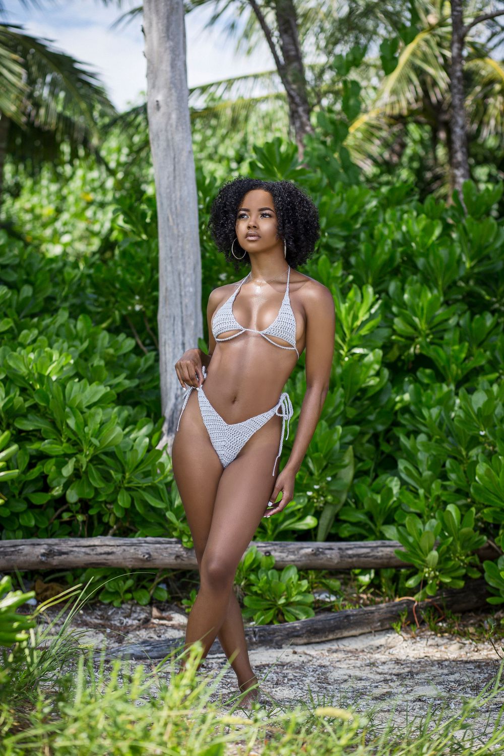 Woman in white crochet bikini standing near palm tree with tropical greenery.