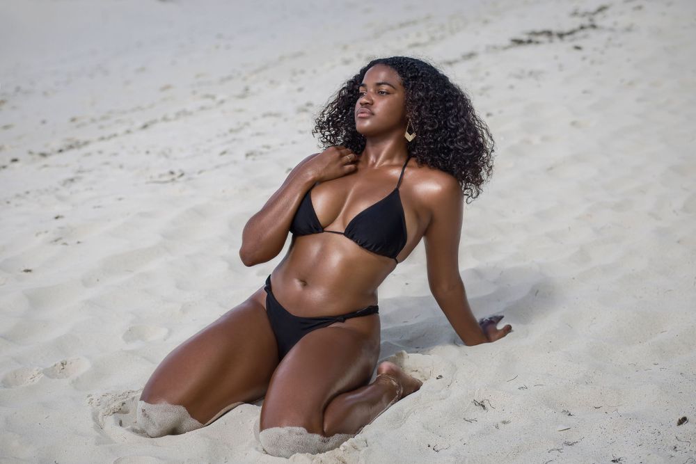 Woman in black bikini sitting on white sand beach with curly hair blowing in the breeze.
