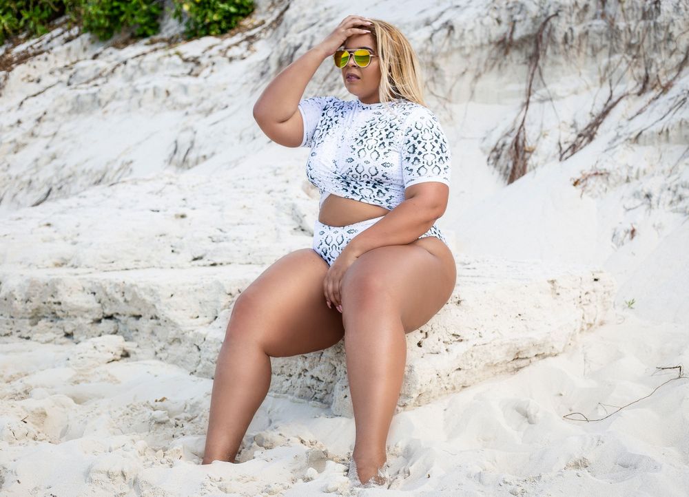 Plus size woman in white floral swimsuit sitting on white sandy beach.