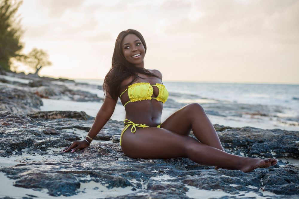 Smiling woman in yellow bikini sitting on rocky shoreline at sunset by the ocean.