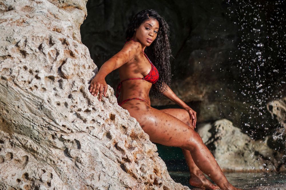 Muscular Black woman in red bikini posing on rocky cliff near waterfall.
