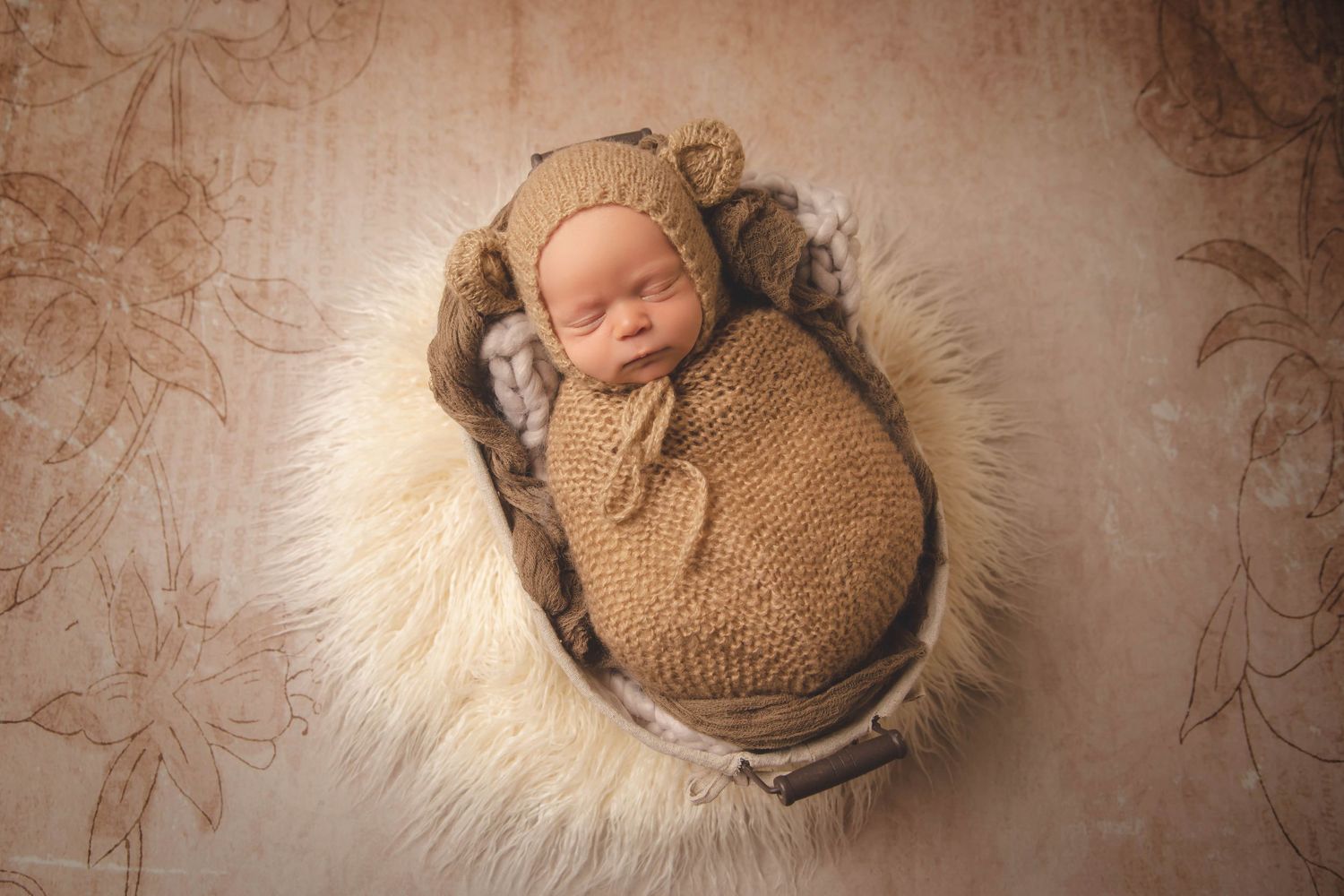 Peaceful sleeping newborn wrapped in brown knit on cream fur backdrop.