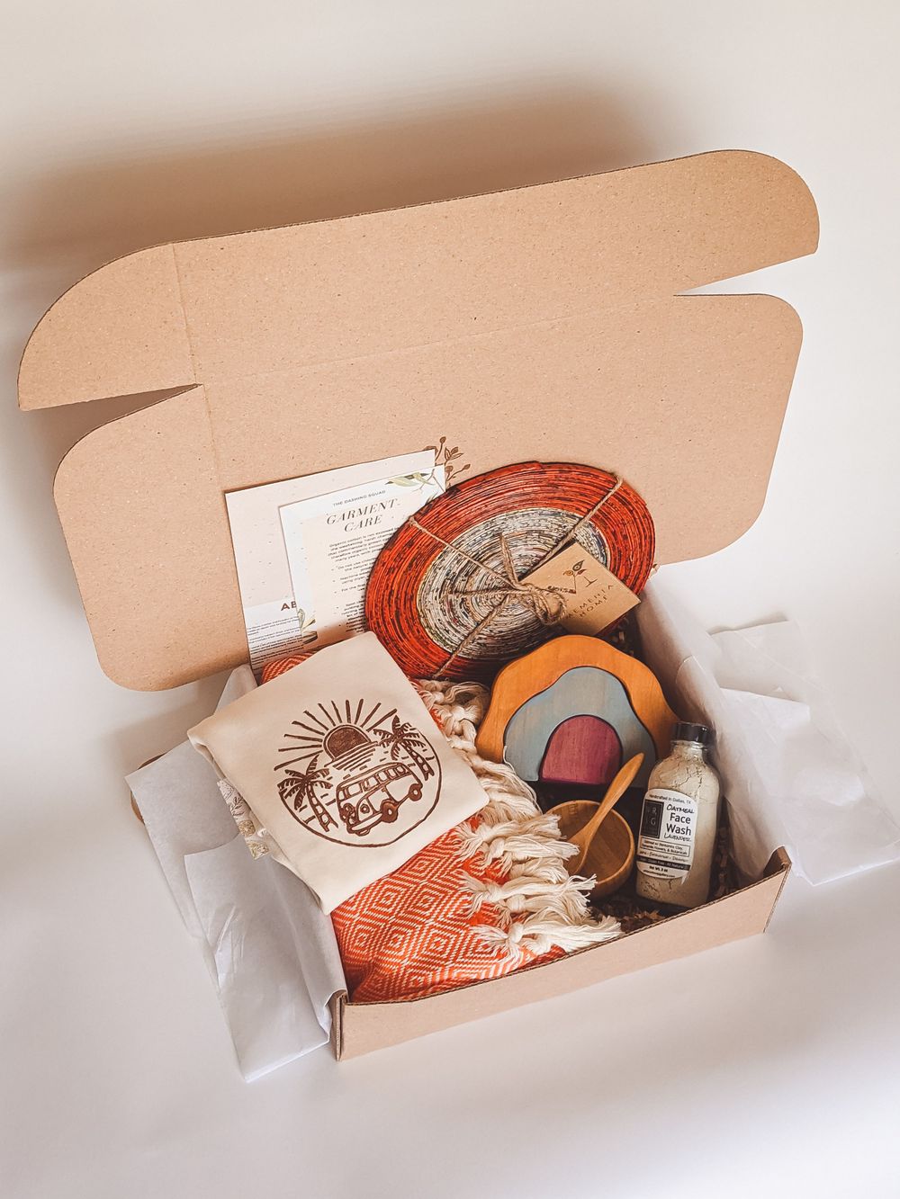 Open gift box containing orange woven coaster, tote bag, small bottle, and assorted items.