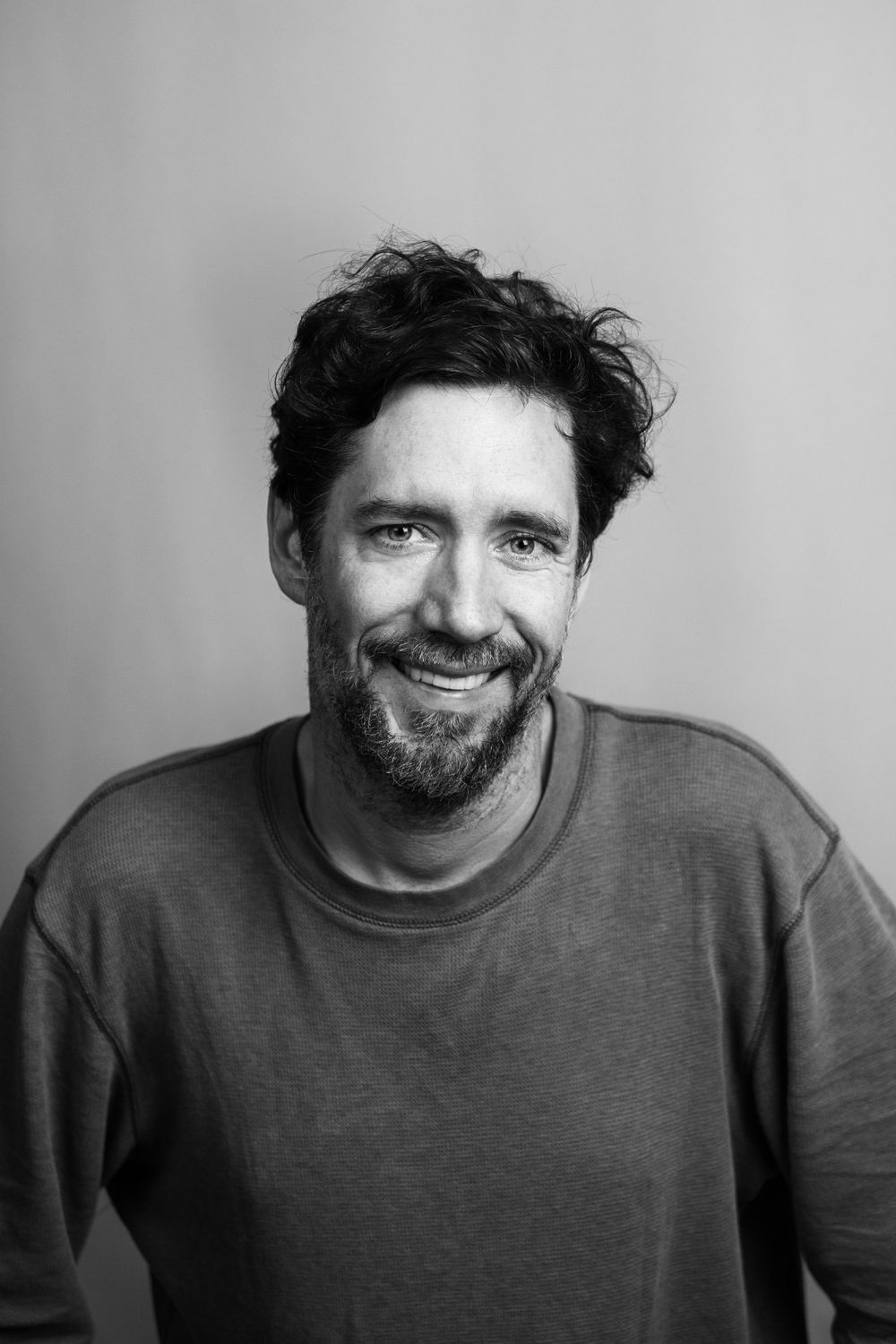 Black and white portrait of smiling bearded man with curly hair wearing a casual sweater.