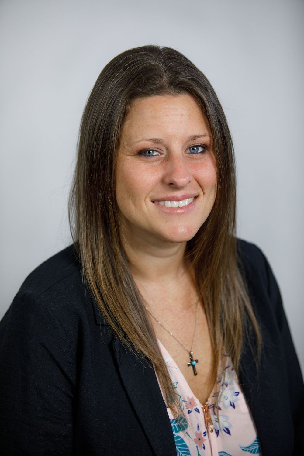 Professional headshot of smiling brunette woman in black blazer and floral blouse on gray background.