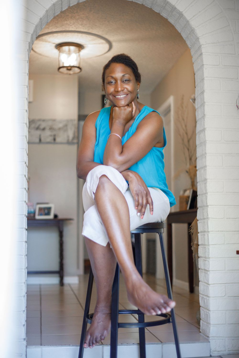 Smiling woman in a blue top and white shorts sitting on a stool in a brick archway at home.