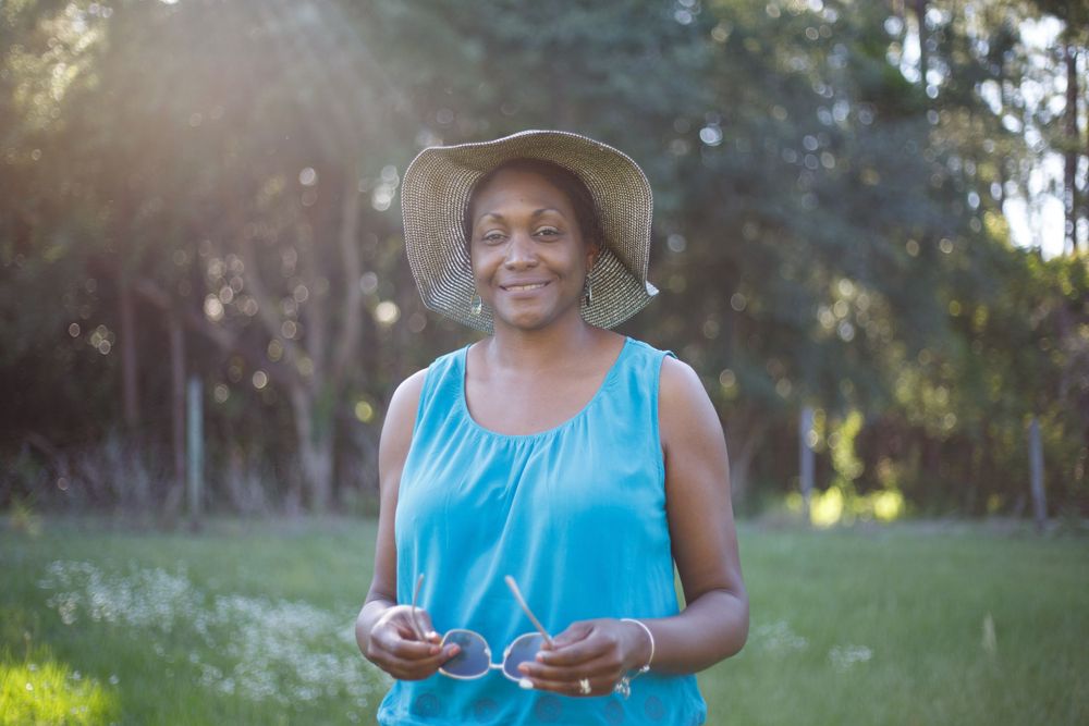 Smiling senior Black woman wearing a straw hat and turquoise dress standing outdoors in sunlit green park.