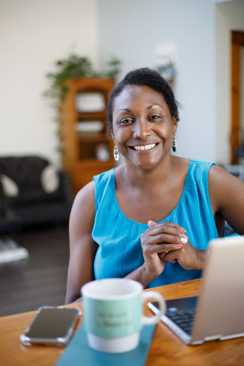 Smiling woman in a blue top sitting at a table with a laptop and coffee mug at home.