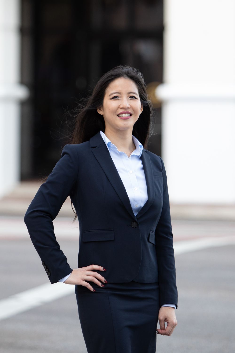 Confident Asian businesswoman in navy suit smiling outdoors with hand on hip.