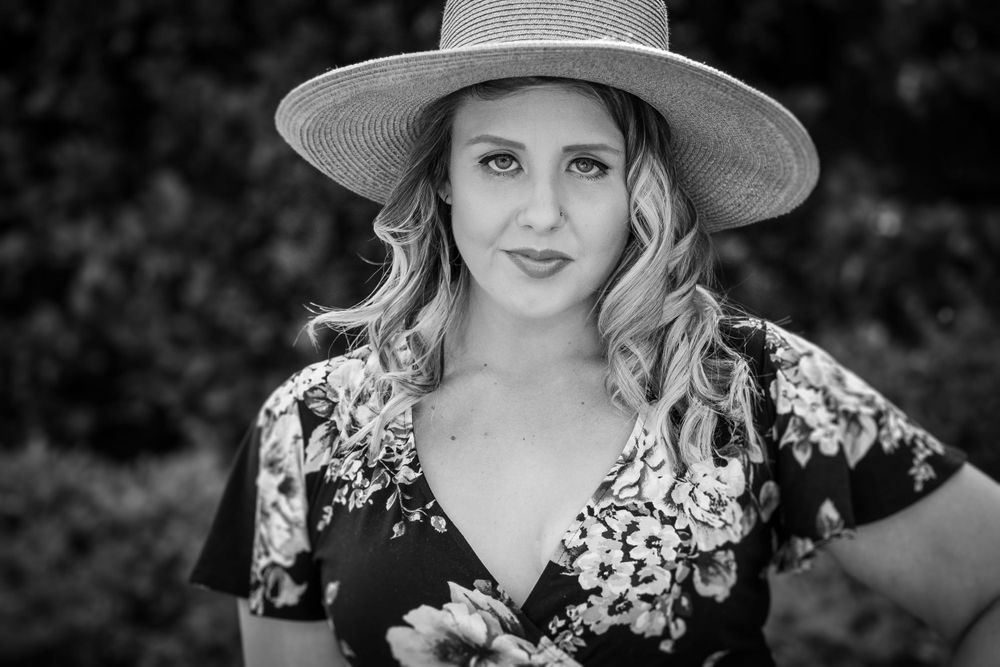 Black and white portrait of a young blonde woman wearing a straw hat and floral dress outdoors.
