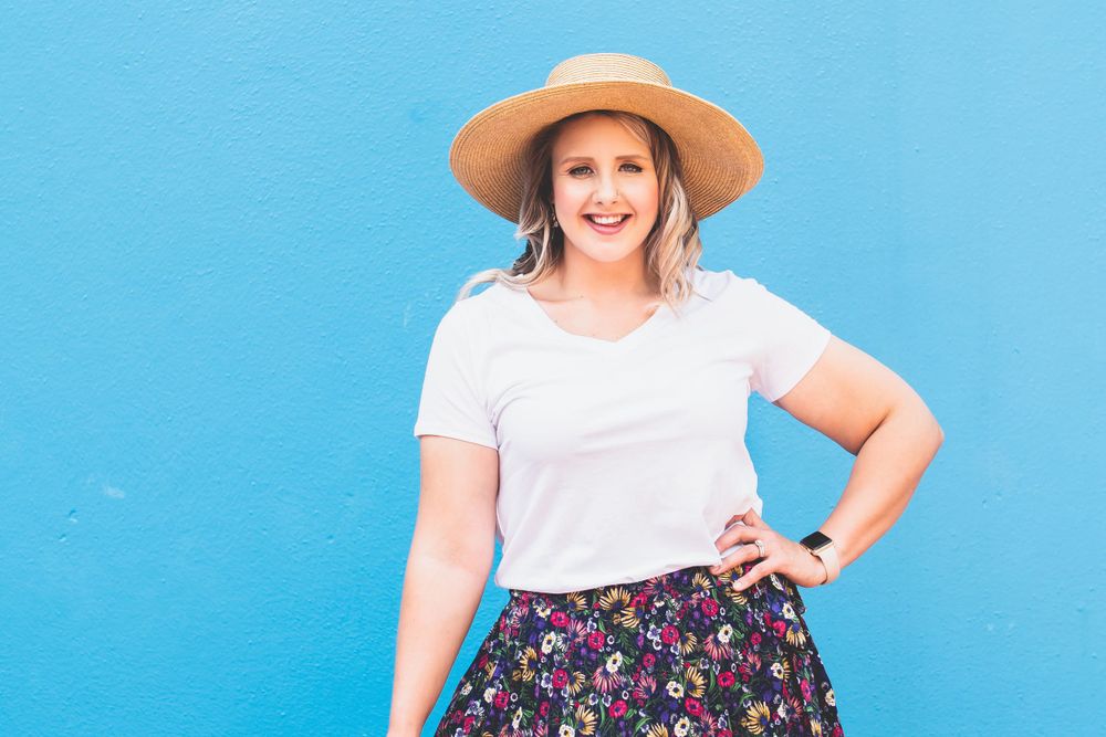 Smiling woman in straw hat, white tee, and floral skirt posing against bright blue wall.