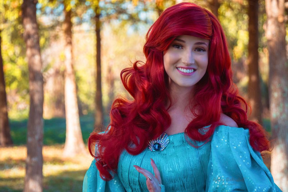 Smiling woman dressed as Ariel from The Little Mermaid with red hair in a teal costume outdoors.