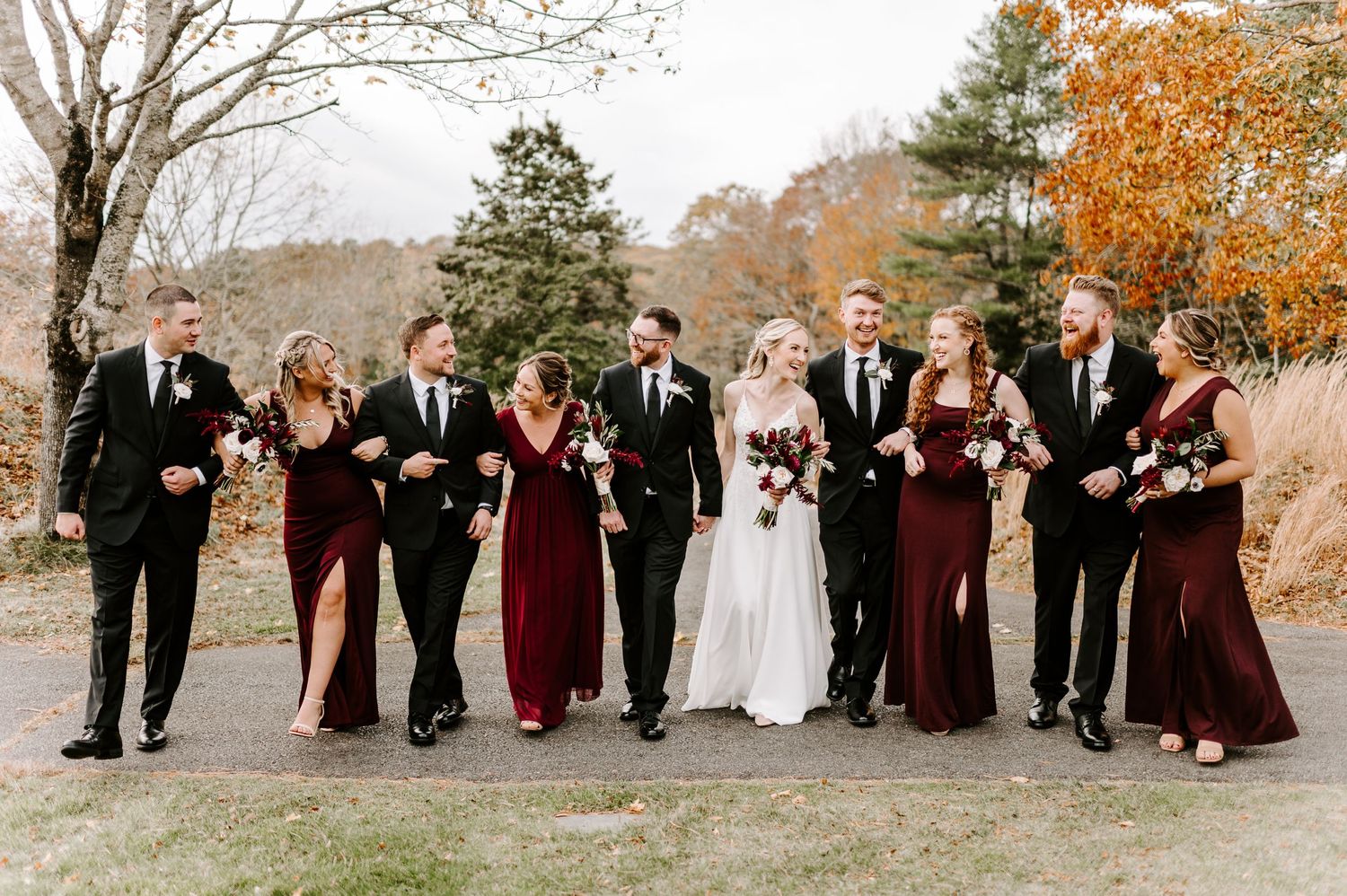Wedding party lines up on path wearing burgundy and black attire amid fall scenery.
