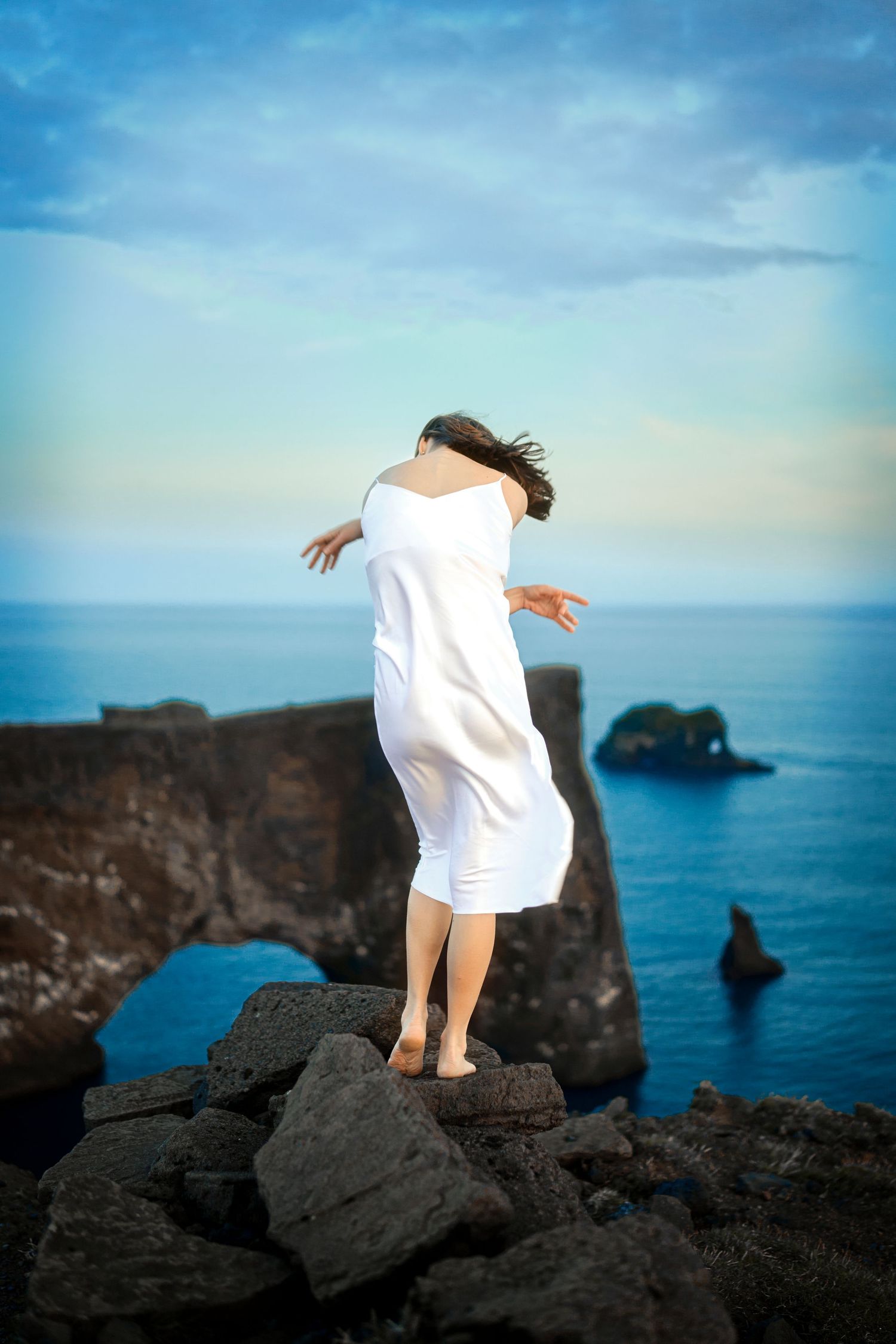 Person in white dress standing on coastal cliff edge overlooking ocean.