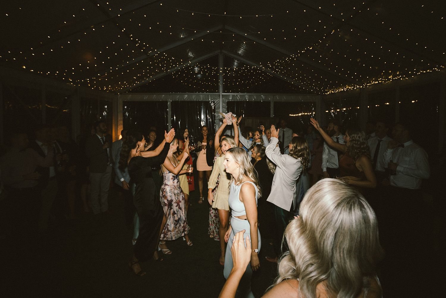 Dark reception venue with wedding guests dancing under romantic string light decor.