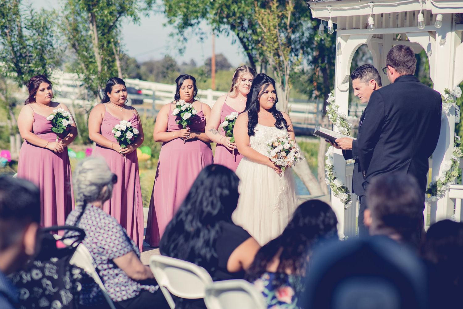 Bride with bridesmaids in pink dresses standing at outdoor wedding altar with white gazebo and officiant.