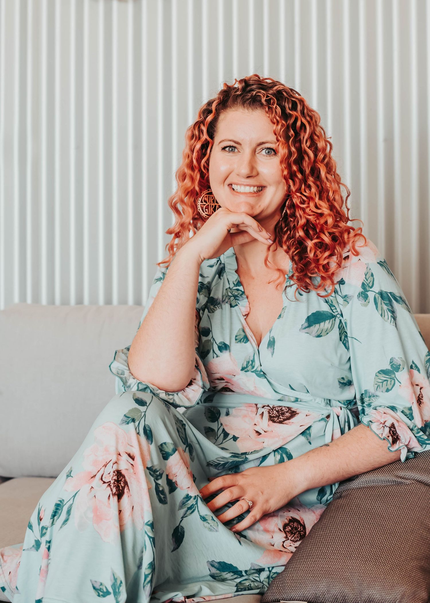 A person in a mint green floral dress with curly copper hair sits casually on a grey couch.