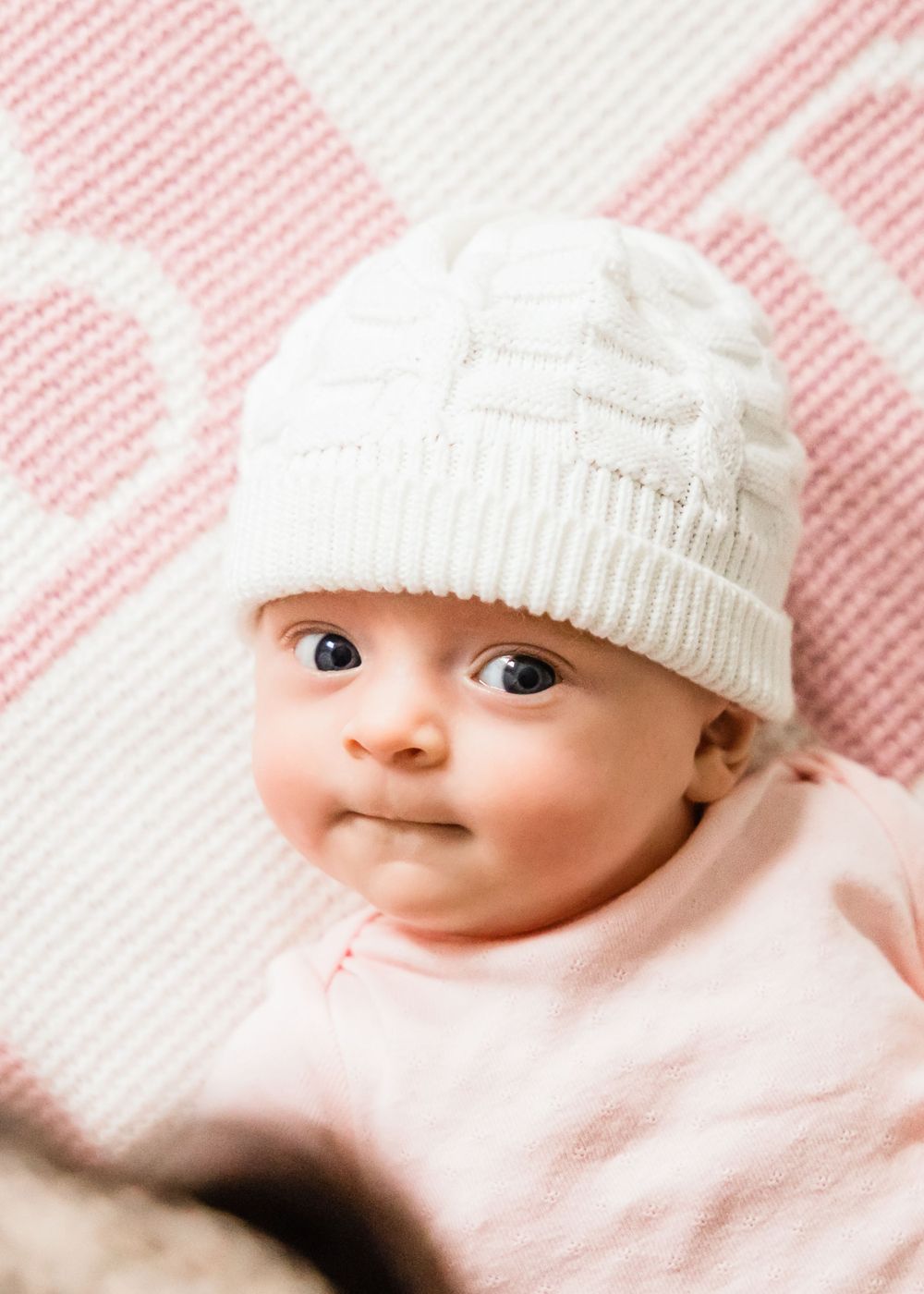 baby girl lifestyle photo on pink blanket with white beanie smiling at camera