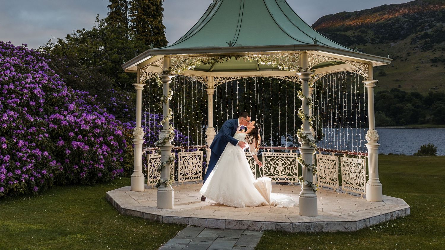 Wedding couple shares intimate moment in illuminated gazebo by lakeside with purple flowers.