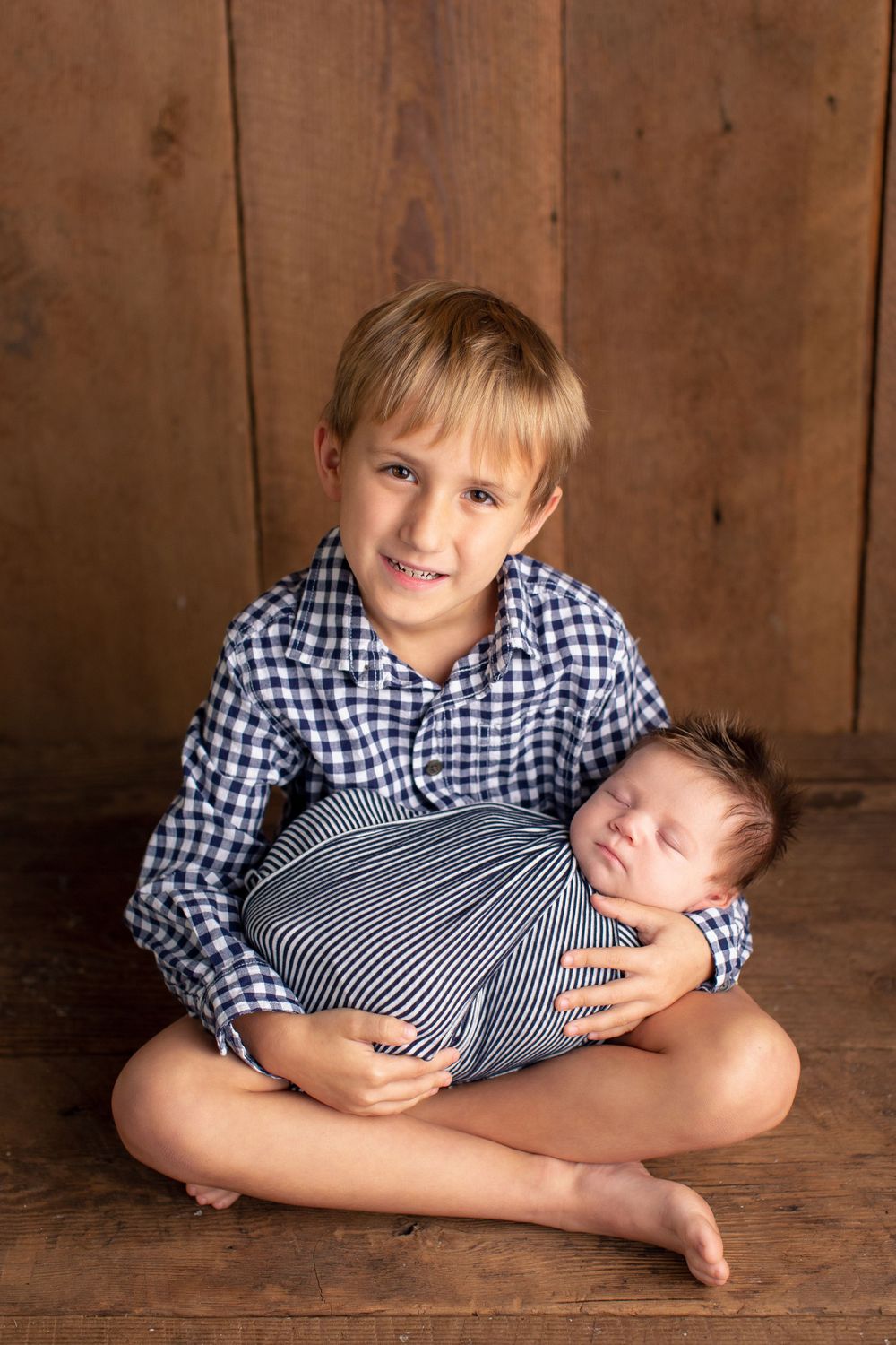 A boy in a checkered shirt holds a swaddled newborn on wooden floor backdrop.