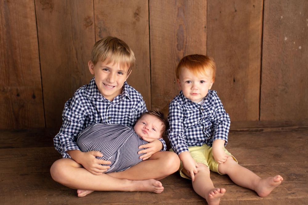 Two siblings in matching blue checkered shirts sit with newborn baby on wooden floor.