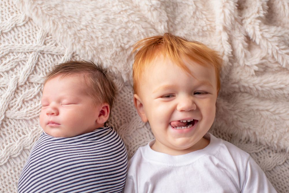 Two siblings lying on white blanket, one sleeping in stripes and one smiling.