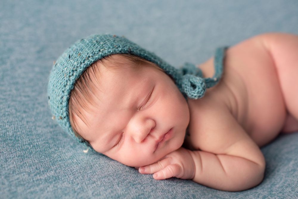 Sleeping newborn posed in teal knit bonnet on blue textured backdrop.