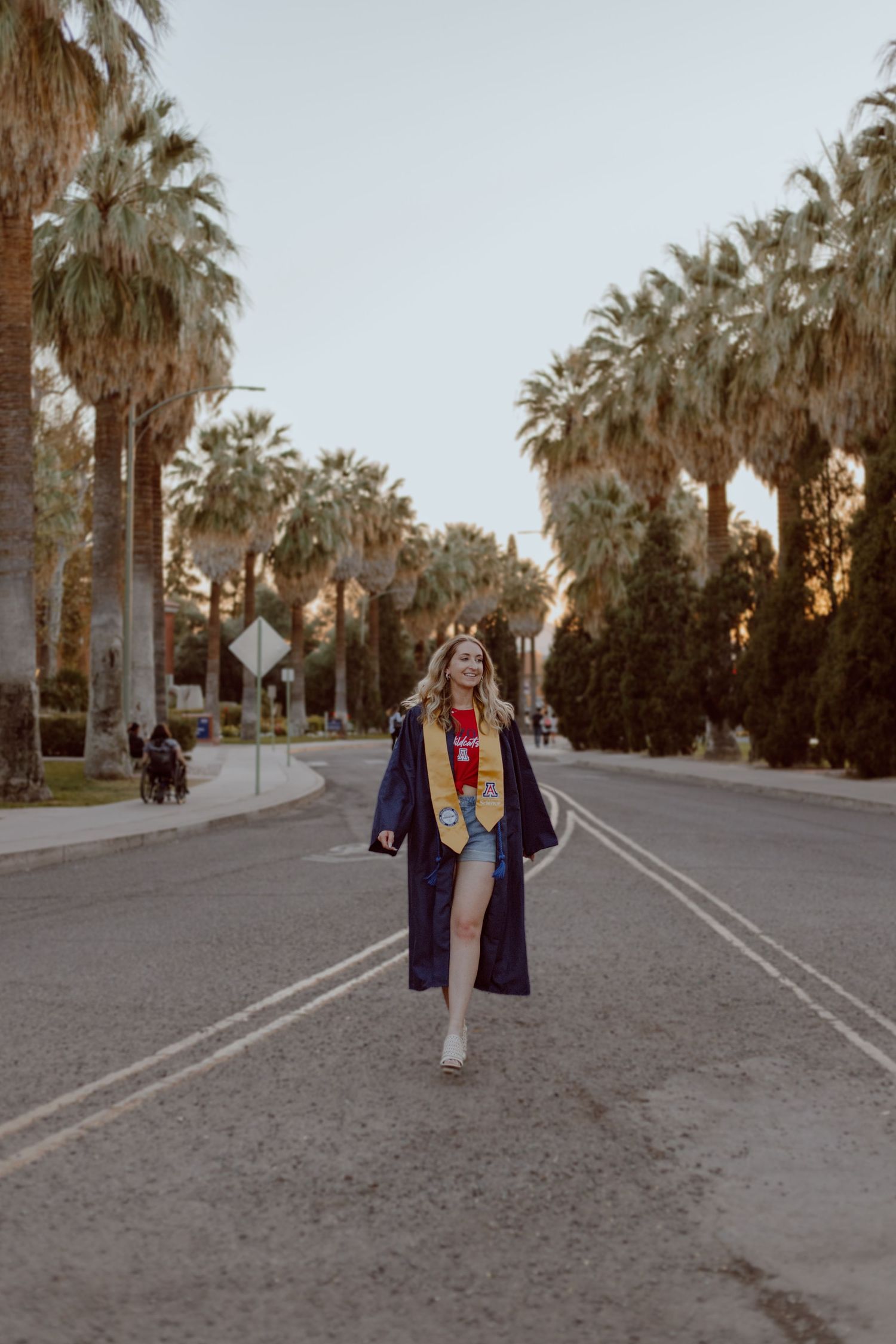 A palm tree-lined path at sunset shows a figure walking in graduation regalia.
