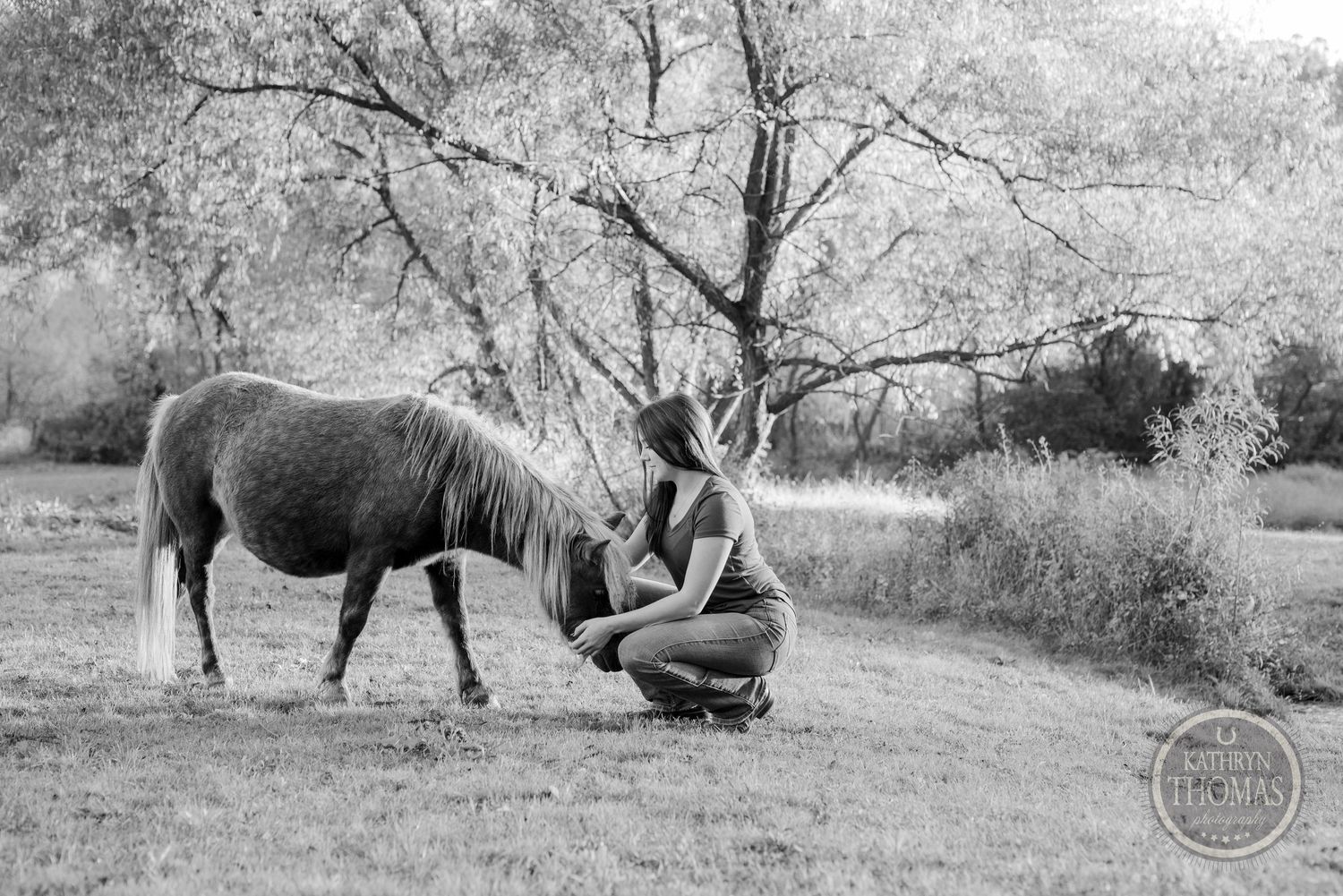 A black and white photo shows a person kneeling beside a grazing pony under a tree.