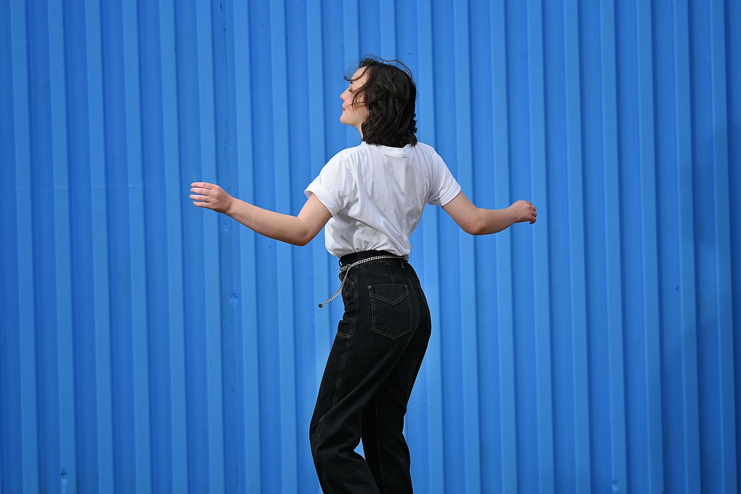Dynamic pose against blue corrugated wall background in casual white t-shirt and black jeans.
