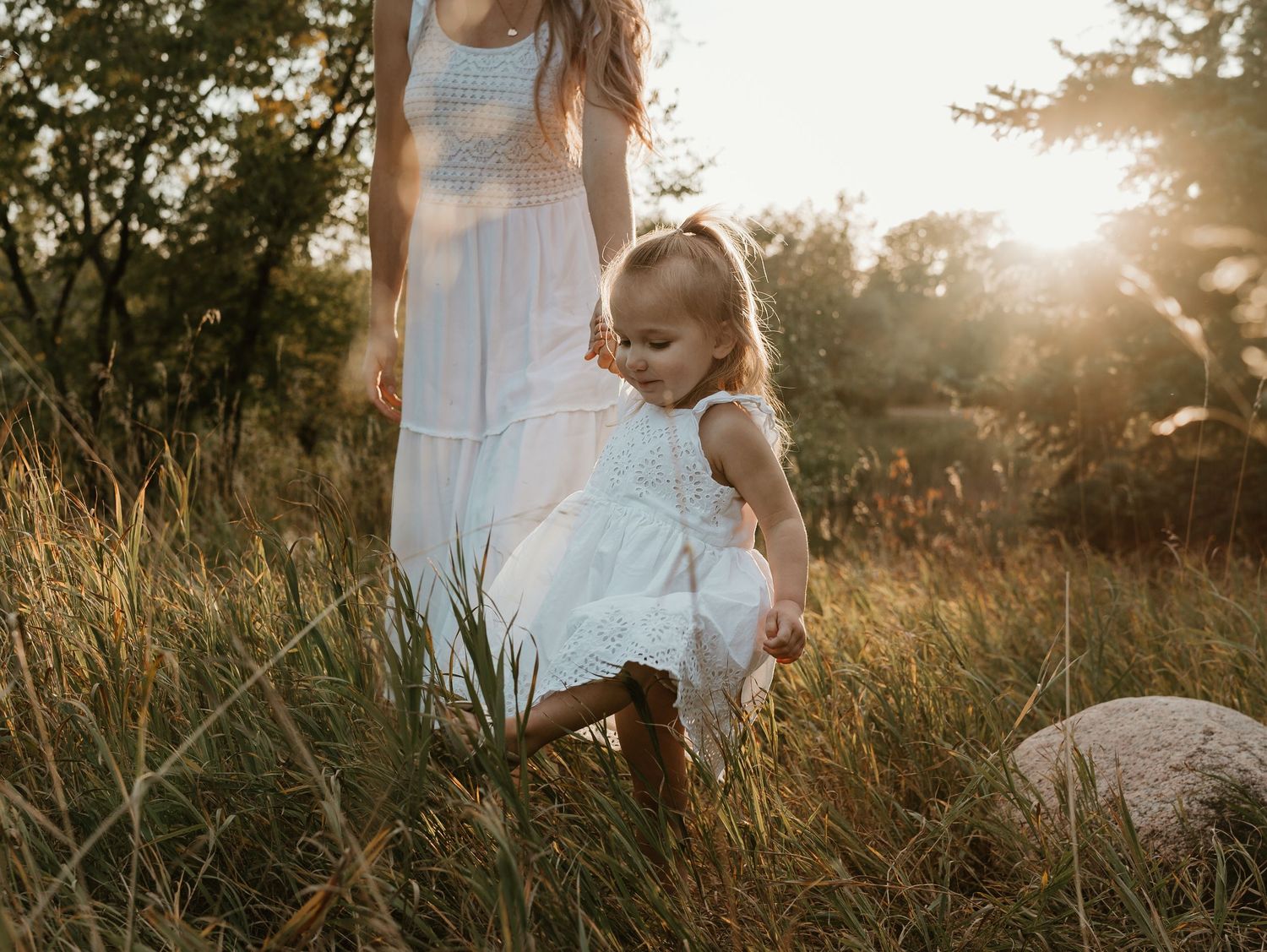 A little girl with blonde hair and a white dress, holding hands with her mom in a golden hour field during sunset in the Okanagan.