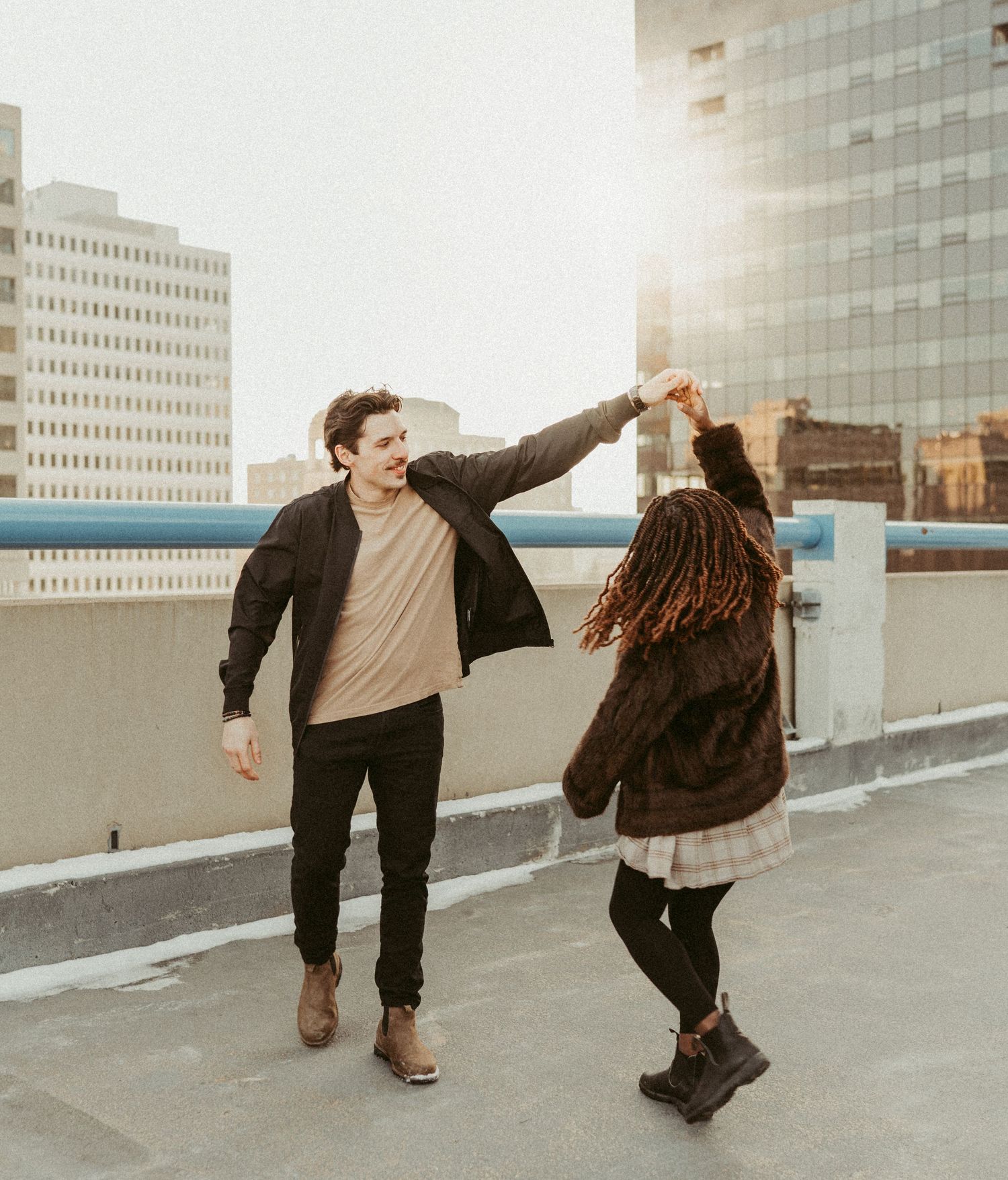 bi racial couple twirling and having fun during their rooftop engagement session with the sun shining through the city scape behind them.