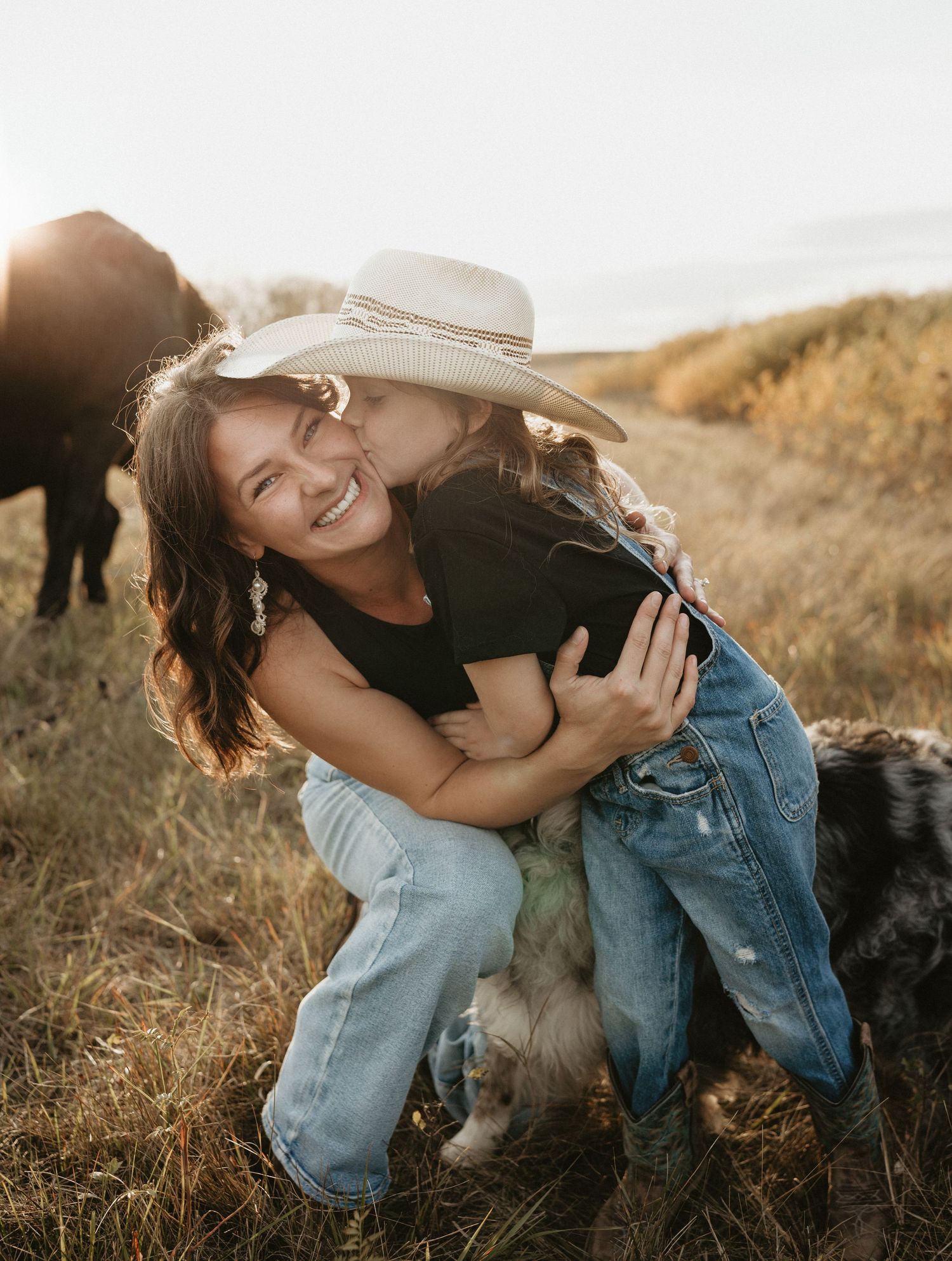 A candid moment of a mother and daughter embracing and laughing in a field during golden hour, while their horse grazes in the background.