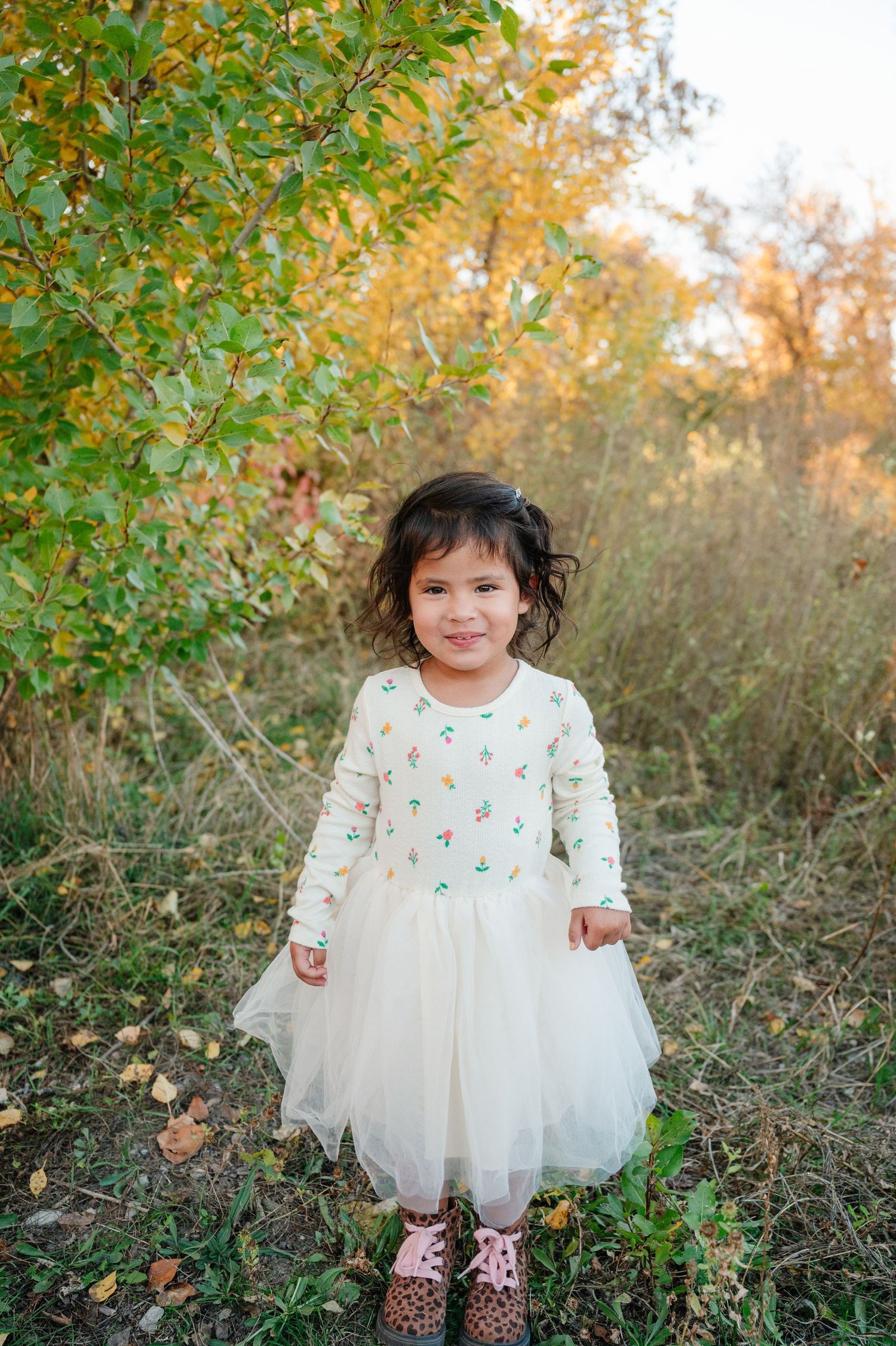 A small child wearing a white tulle dress poses outdoors among autumn foliage.