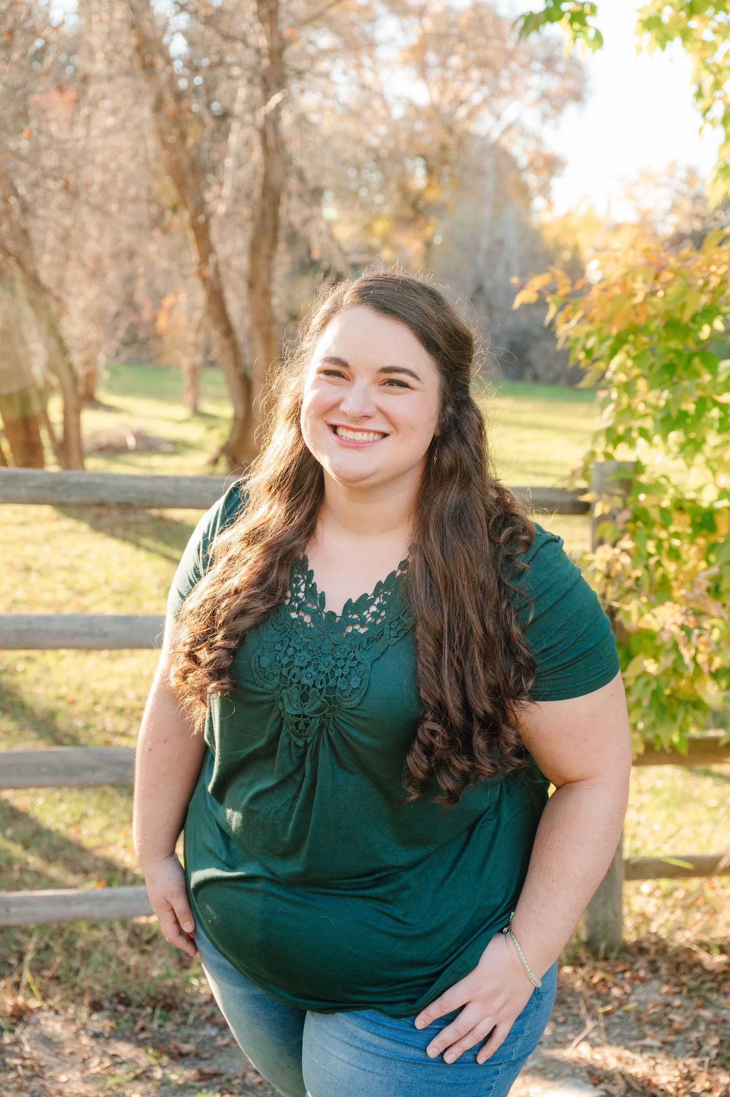 Portrait of woman in green top smiling outdoors with autumn scenery in background.