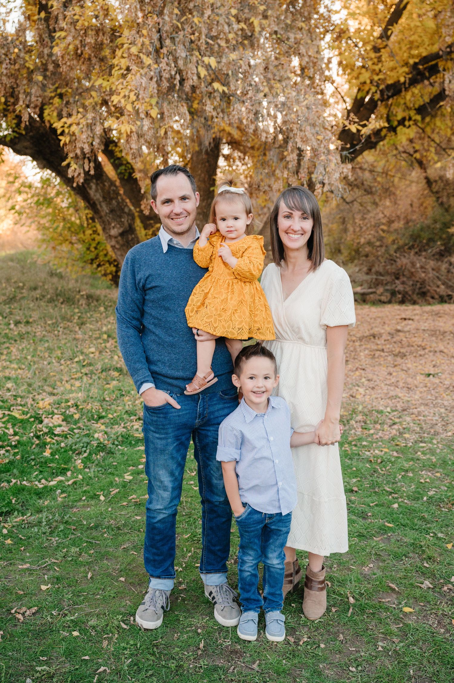 Family of four poses together under golden autumn trees in outdoor setting.