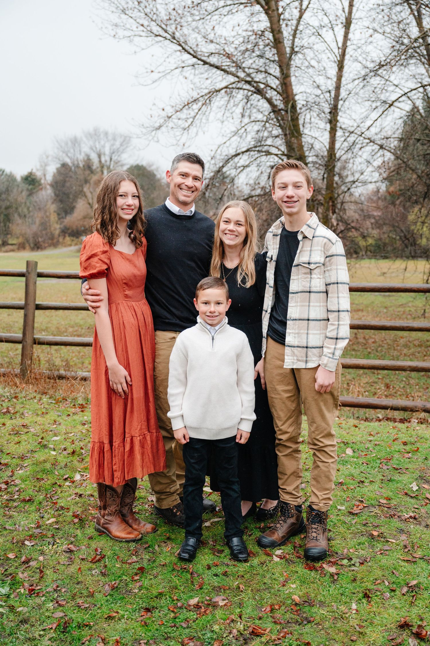 Family of five poses together in front of rustic wooden fence during fall season.
