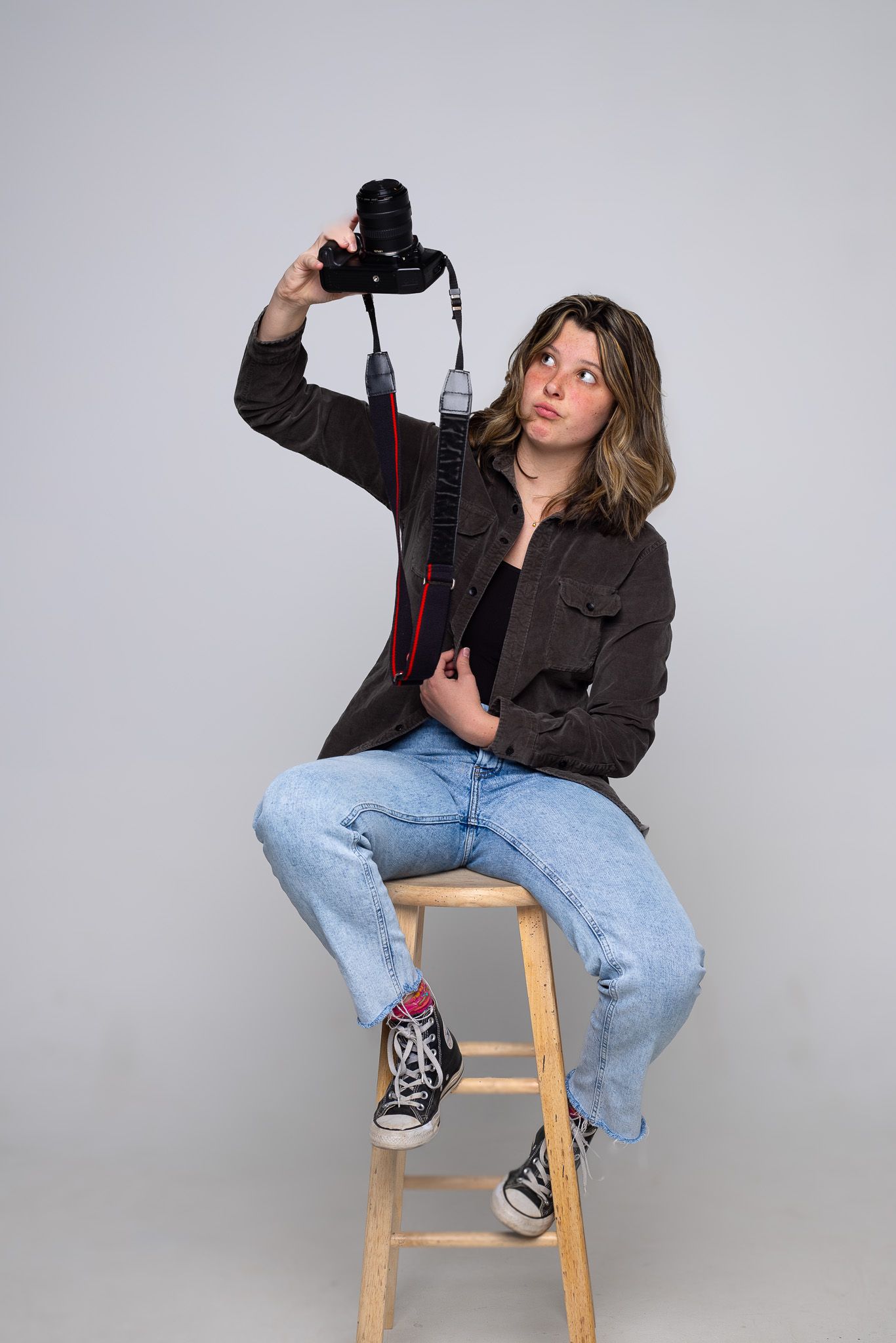 Photographer in casual attire holding camera while sitting on wooden stool in studio setting.