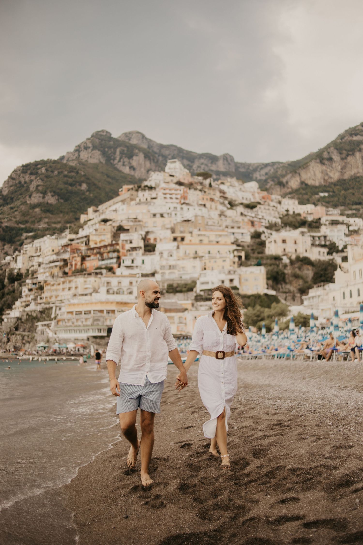 Couple running joyfully on beach with Italian coastal village of Positano cascading down cliffs behind them.