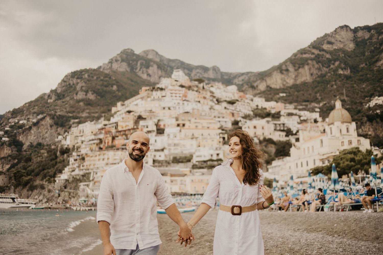 Romantic couple holding hands on Positano beach with stunning Amalfi Coast village backdrop.