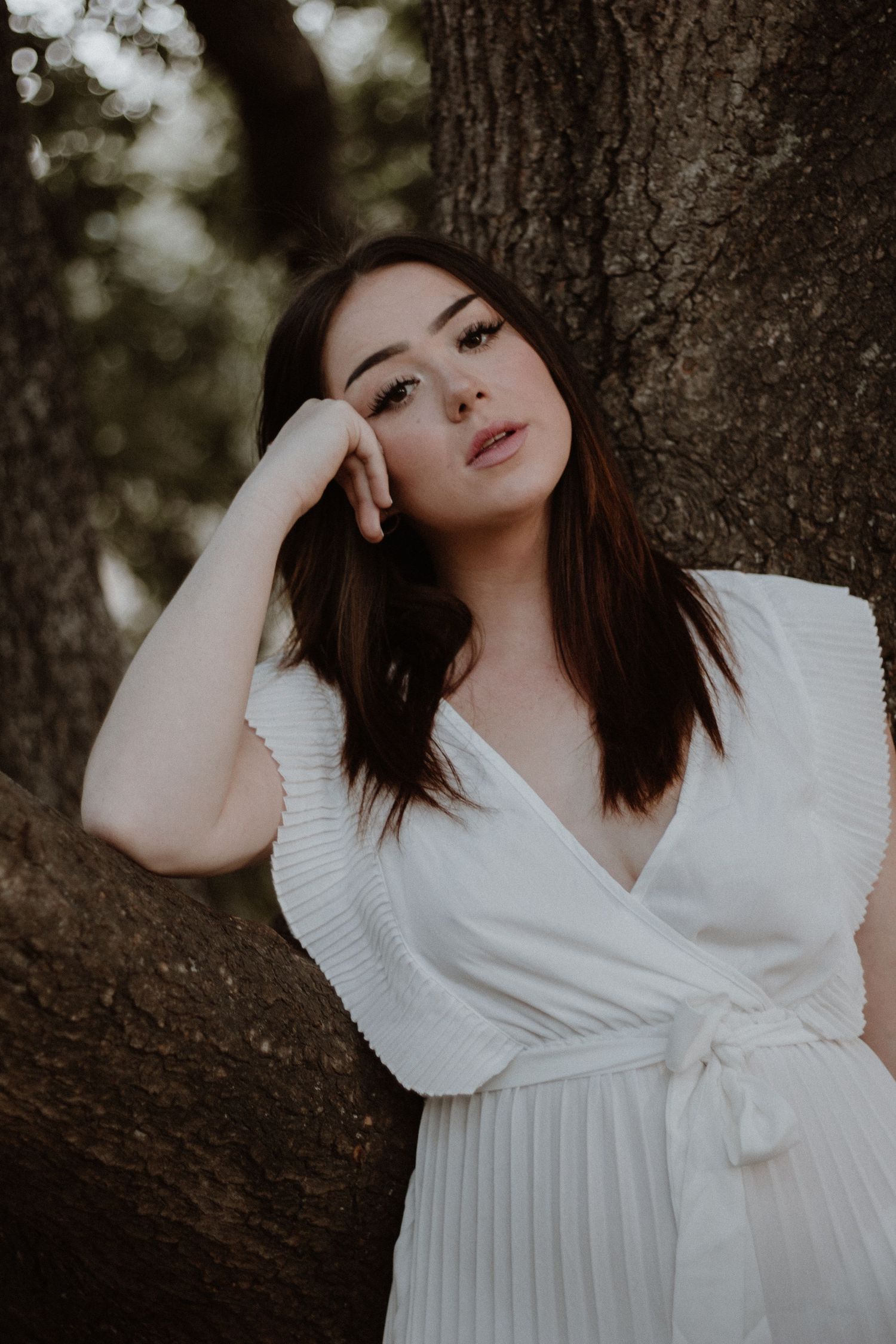 Artistic portrait of a woman in a white pleated dress leaning against a tree trunk.