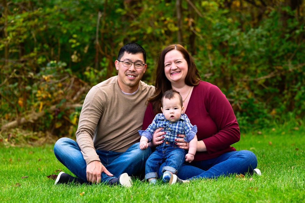 A young family sitting together on grass with autumn foliage in the background.