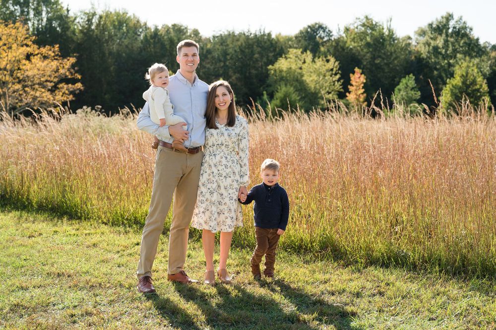 Family standing together in a golden field at sunset with tall grass and trees in the background.